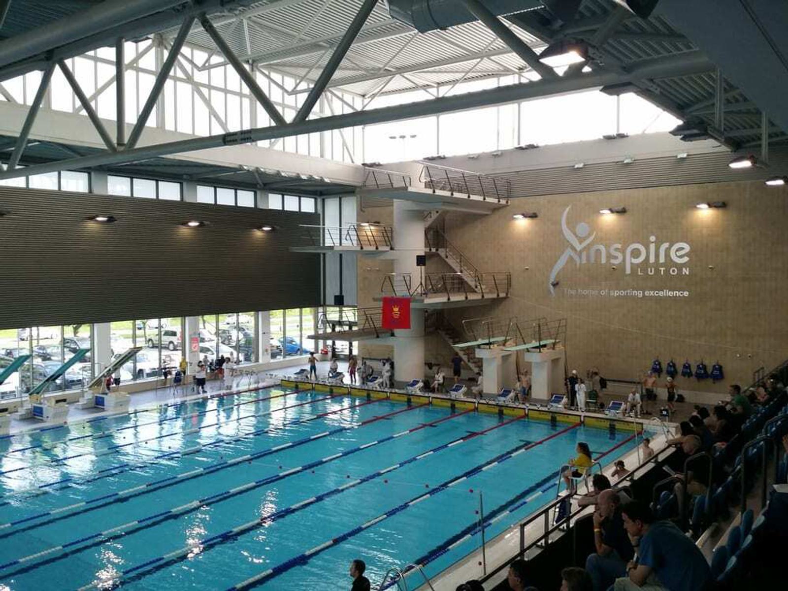 Indoor swimming pool with lanes and diving platforms. Spectators watch from the stands. A large logo on the wall reads "Inspire Luton: The home of sporting excellence." Bright, spacious area with natural light.