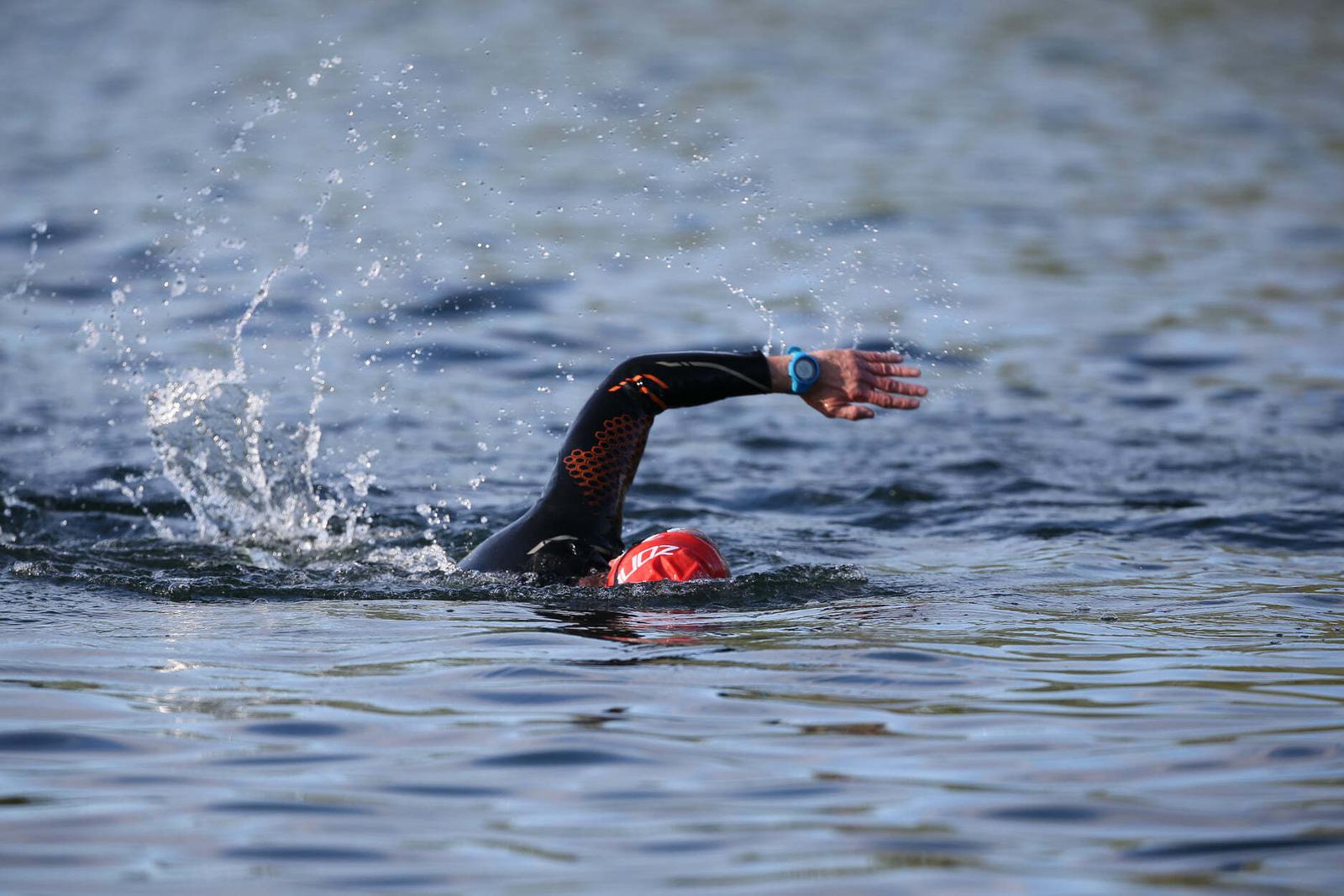 A swimmer wearing a black wetsuit and red swim cap is seen performing a freestyle stroke in open water. Ripples and splashes surround them as they move through the blue surface.