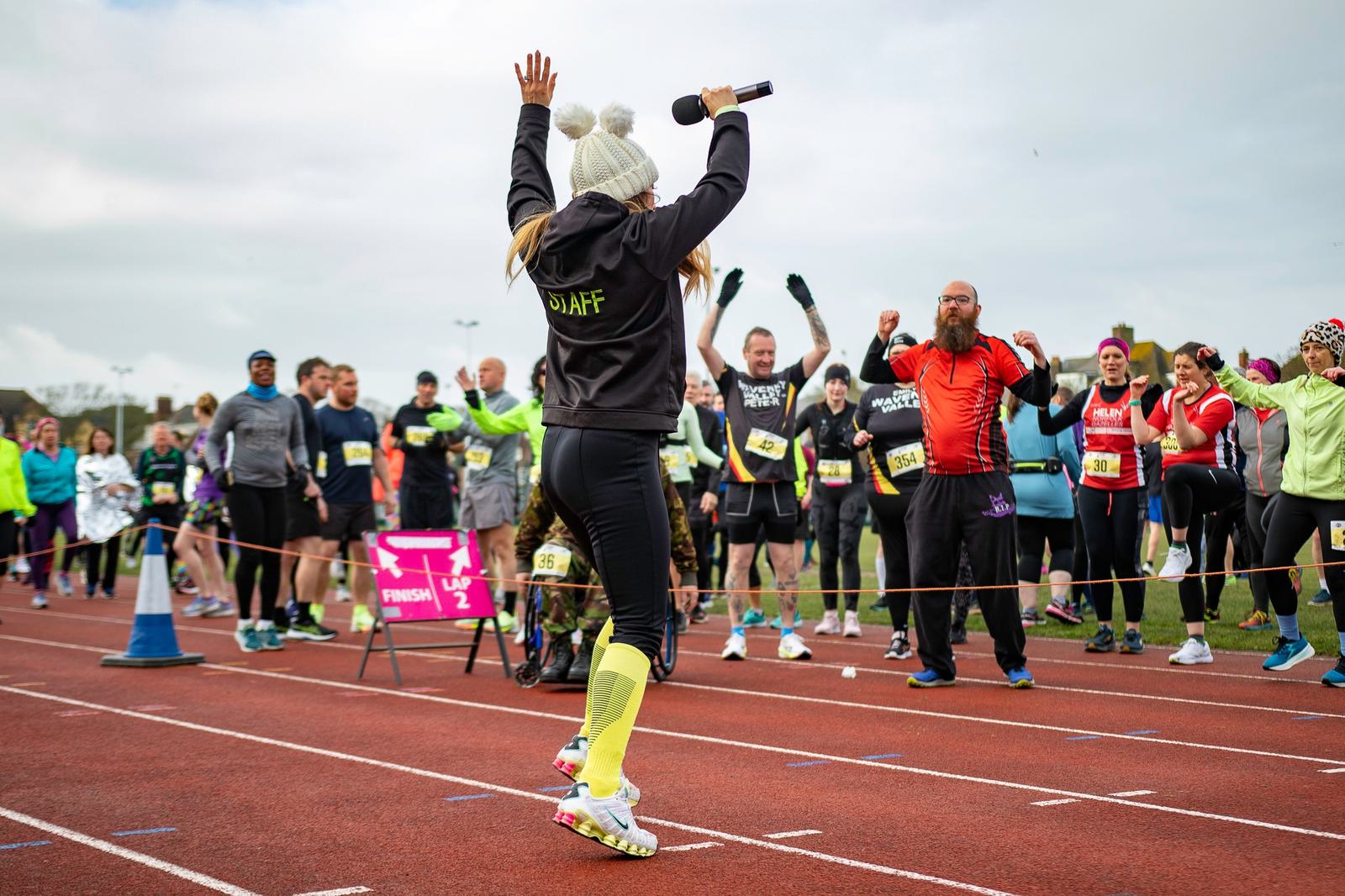 A race facilitator, wearing a beanie and "STAFF" jacket, energetically raises a hand at a starting line on a track. A crowd of runners waits behind, stretching and preparing to start. A pink sign marks "FINISH" in the background.
