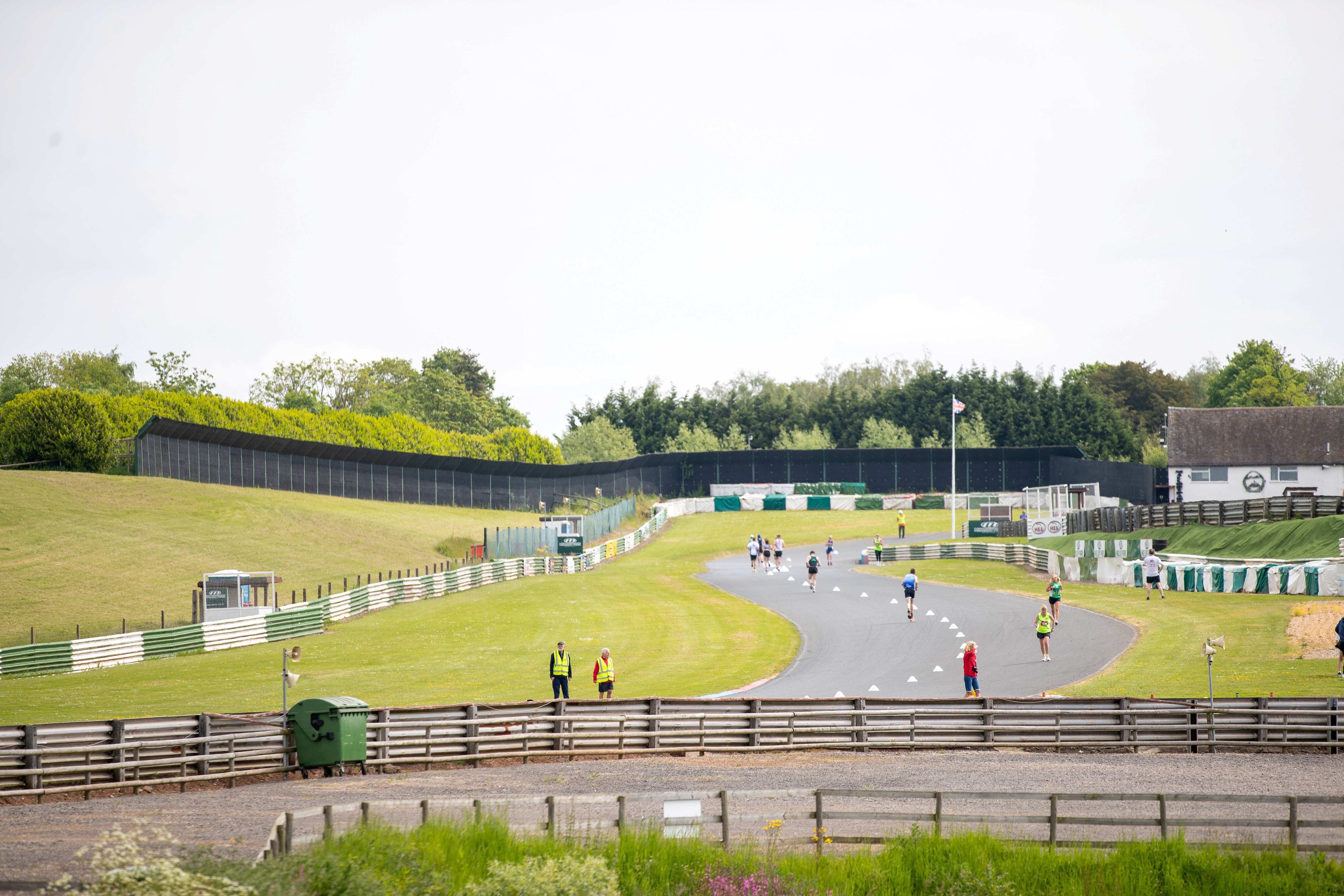People walking on a curved race track surrounded by green grass and trees. Some are wearing high-visibility vests. A barrier and a white building with a dark roof are visible on the right. Overcast sky in the background.