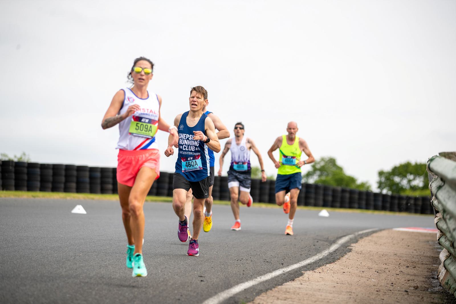 Four runners participating in a race on a paved road. The lead runner is wearing sunglasses, a white top, and pink shorts. Others follow closely, wearing athletic gear. The background features tires and vegetation under an overcast sky.