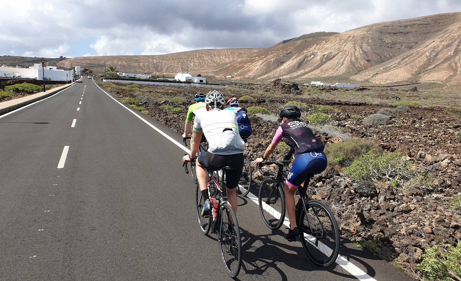 Four cyclists ride on a paved road through a rocky, mountainous landscape under a partly cloudy sky, with white buildings in the distance.