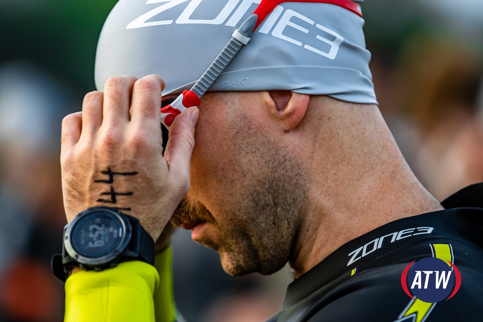 A person wearing a gray swim cap with "ZONE3" printed on it adjusts their red goggles. The individual has a mustache, is wearing a black wetsuit, and has a sports watch on their wrist. The back of their hand has numbers written on it. The ATW logo is visible in the corner.