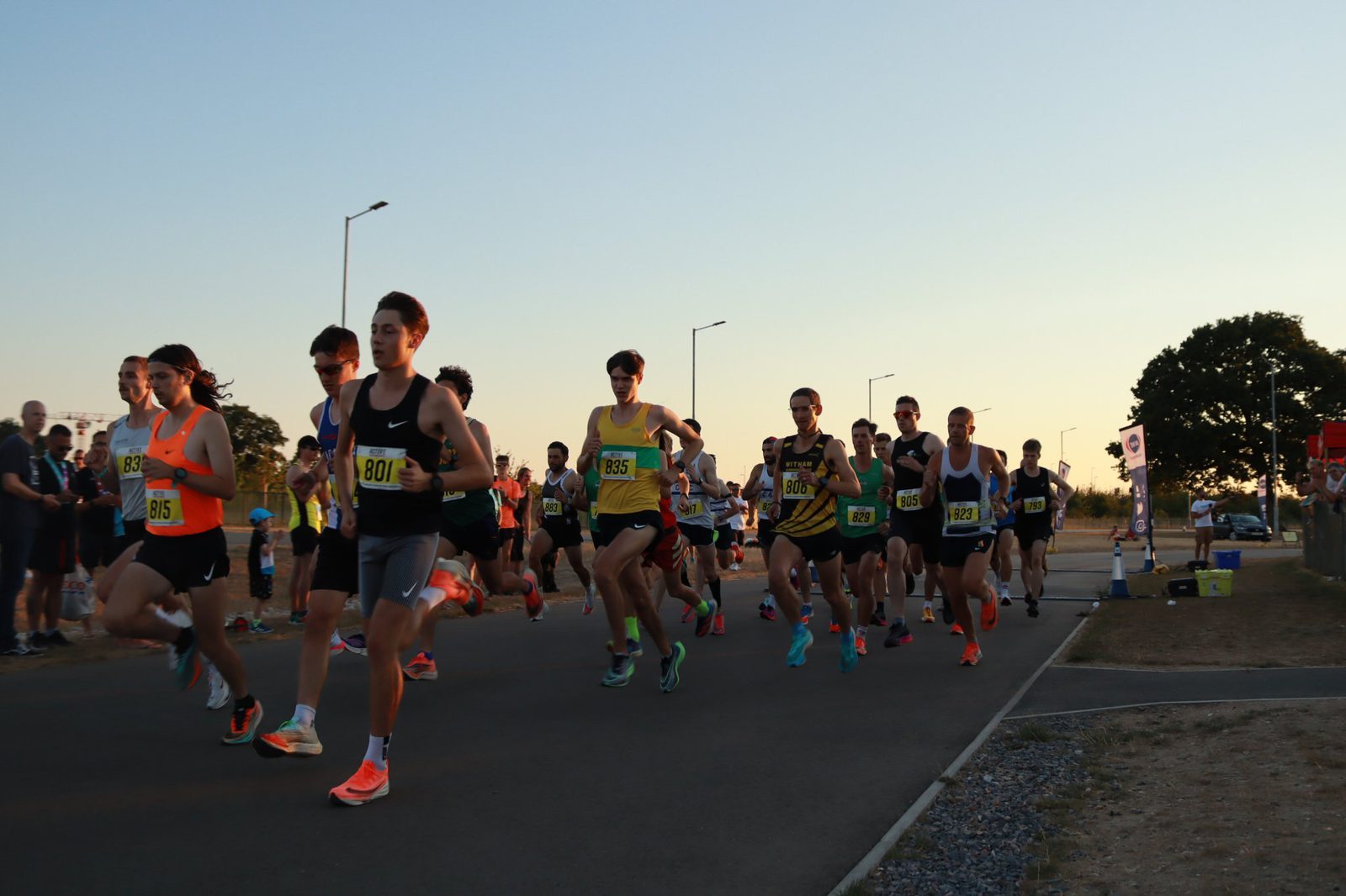 A group of runners competing in a road race, wearing various athletic attire and numbered bibs, sprinting along a paved surface with spectators visible in the background. The setting appears to be during either early morning or late afternoon, indicated by the low sunlight.