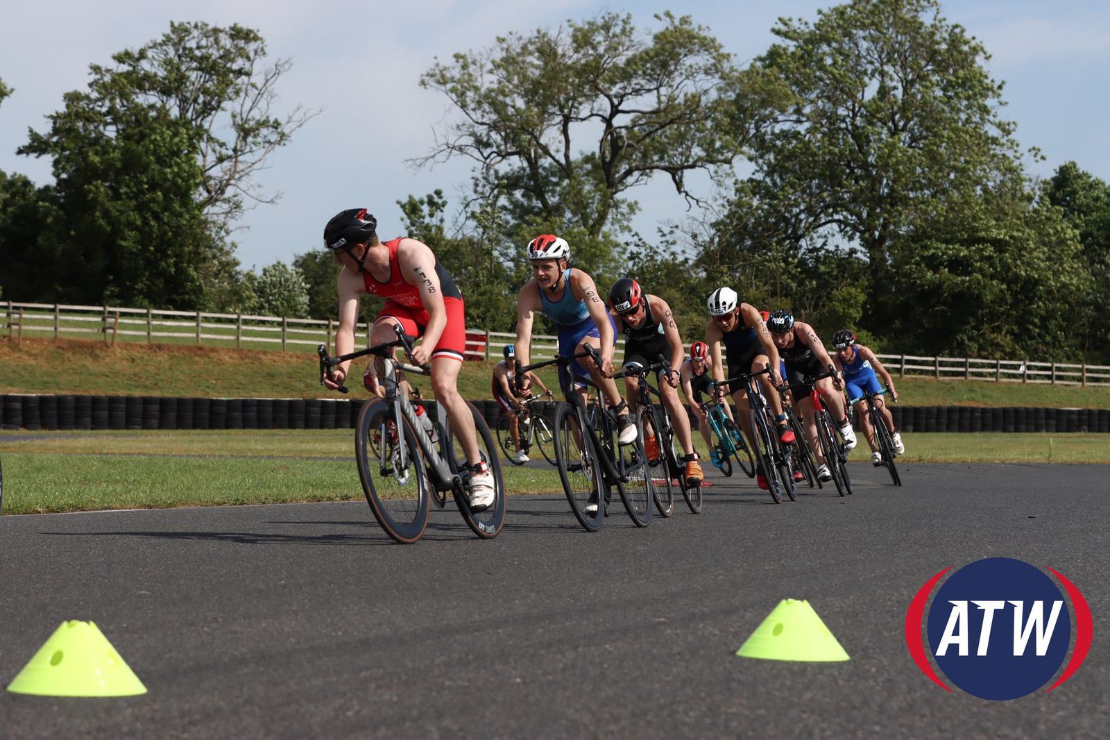 Cyclists in a race on a paved track, with several cones marking the course. The riders are wearing helmets and are in close formation. Trees and a fence line the background, and a "ATW" logo is in the foreground.