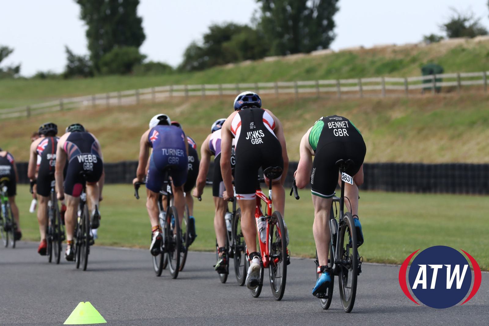 Cyclists in a race on a paved track, wearing helmets and colorful jerseys with names or initials and "GBR" printed on them. Trees and a wooden fence line the grassy surroundings. An "ATW" logo is in the bottom right corner.
