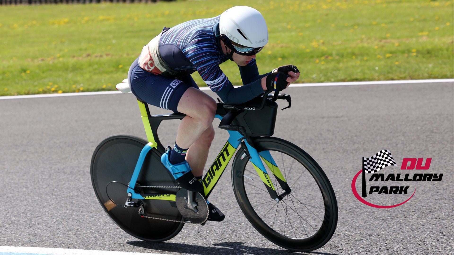A cyclist wearing a blue helmet and striped outfit rides a blue Giant time trial bike on a racetrack. The logo "Mallory Park" with a checkered flag is visible on the right.