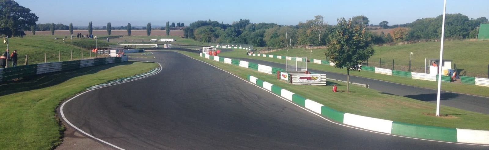 A winding race track with green and white curbs snakes through a grassy landscape under a clear blue sky. Trees and hills are visible in the background. The track is fenced, and there are a few people scattered around the area, possibly officials or spectators.