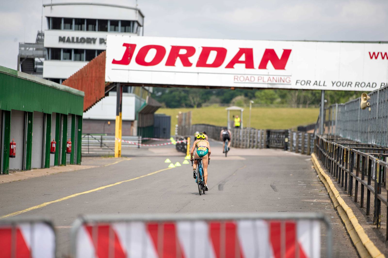 Two cyclists in a race head towards a banner that reads "JORDAN Road Planing" at a cycling track. One cyclist follows closely behind the other on a paved track bordered by green buildings on the left and metal barriers on the right. It is a cloudy day.