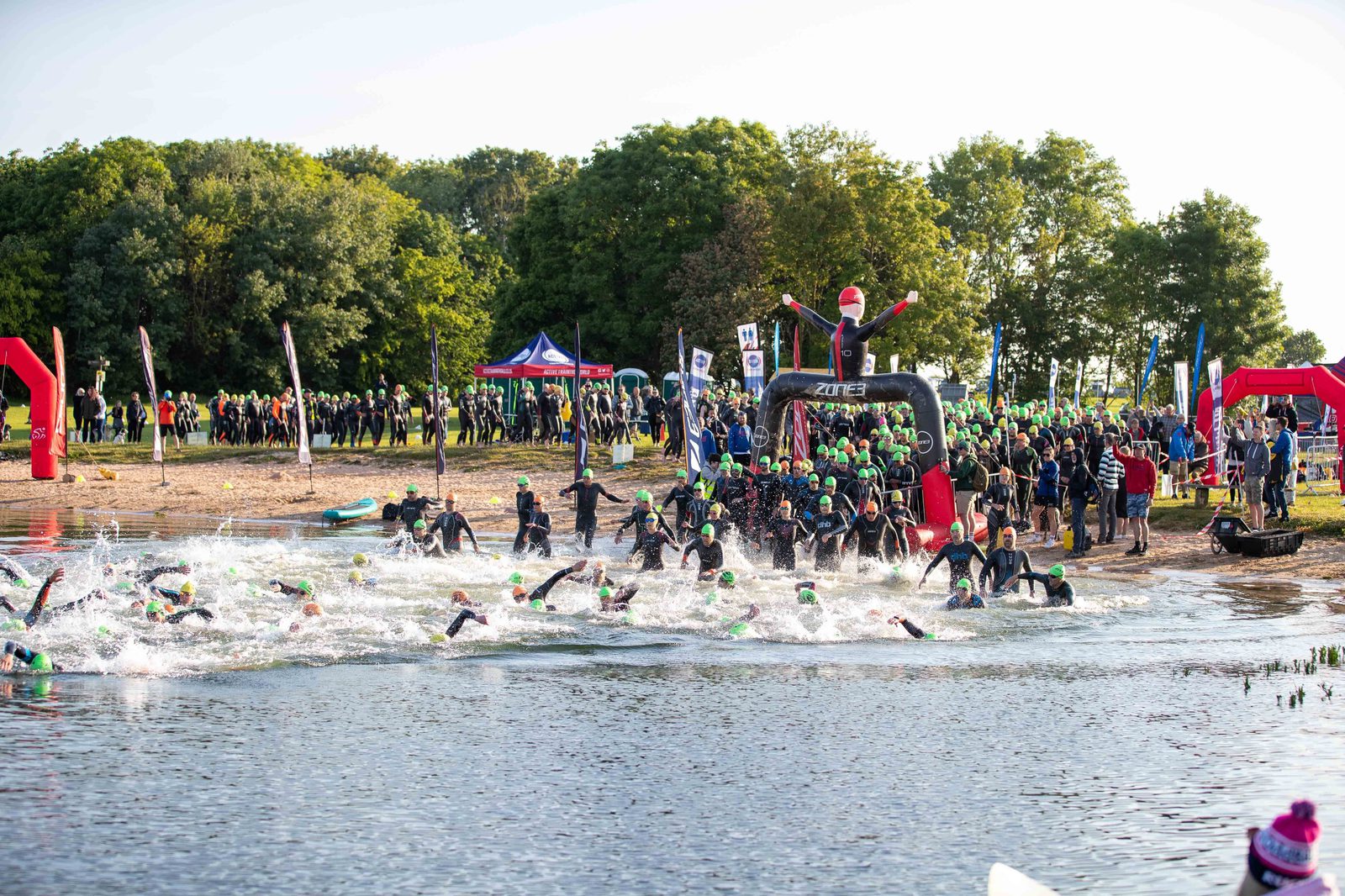 A large group of triathletes wearing wetsuits and green swim caps start the swimming portion of a triathlon by running into a lake. Spectators gather along the shore, with event banners and a timing arch in the background. The scene is lively and full of energy.