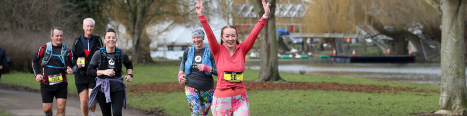 A group of runners jogs along a path near a lake on an overcast day. The runner in the foreground, wearing a pink top and colorful leggings, raises her arms in celebration. The other runners, dressed in various athletic gear, smile as they follow along the route.