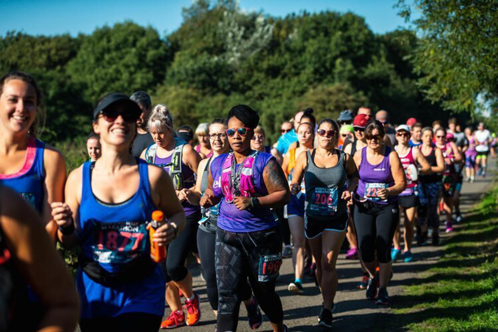 A large group of runners, mostly women, participate in an outdoor race on a sunny day. They are wearing race bibs and a variety of athletic gear, including tank tops, hats, and sunglasses. Trees and greenery line the path they're running on.