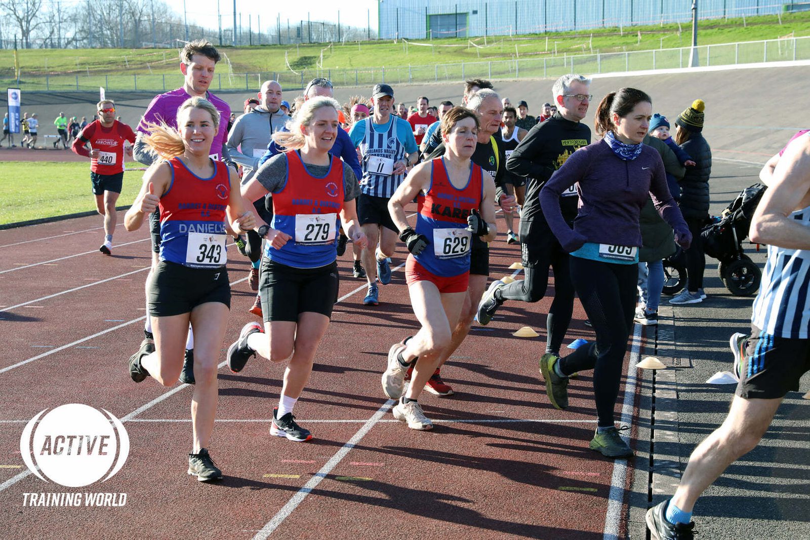 A group of runners, including women wearing numbered bibs, are participating in an outdoor race. Some runners are in red shirts with numbers 343, 279, and 22. It's a sunny day at a stadium track. The "Active Training World" logo is visible in the corner.