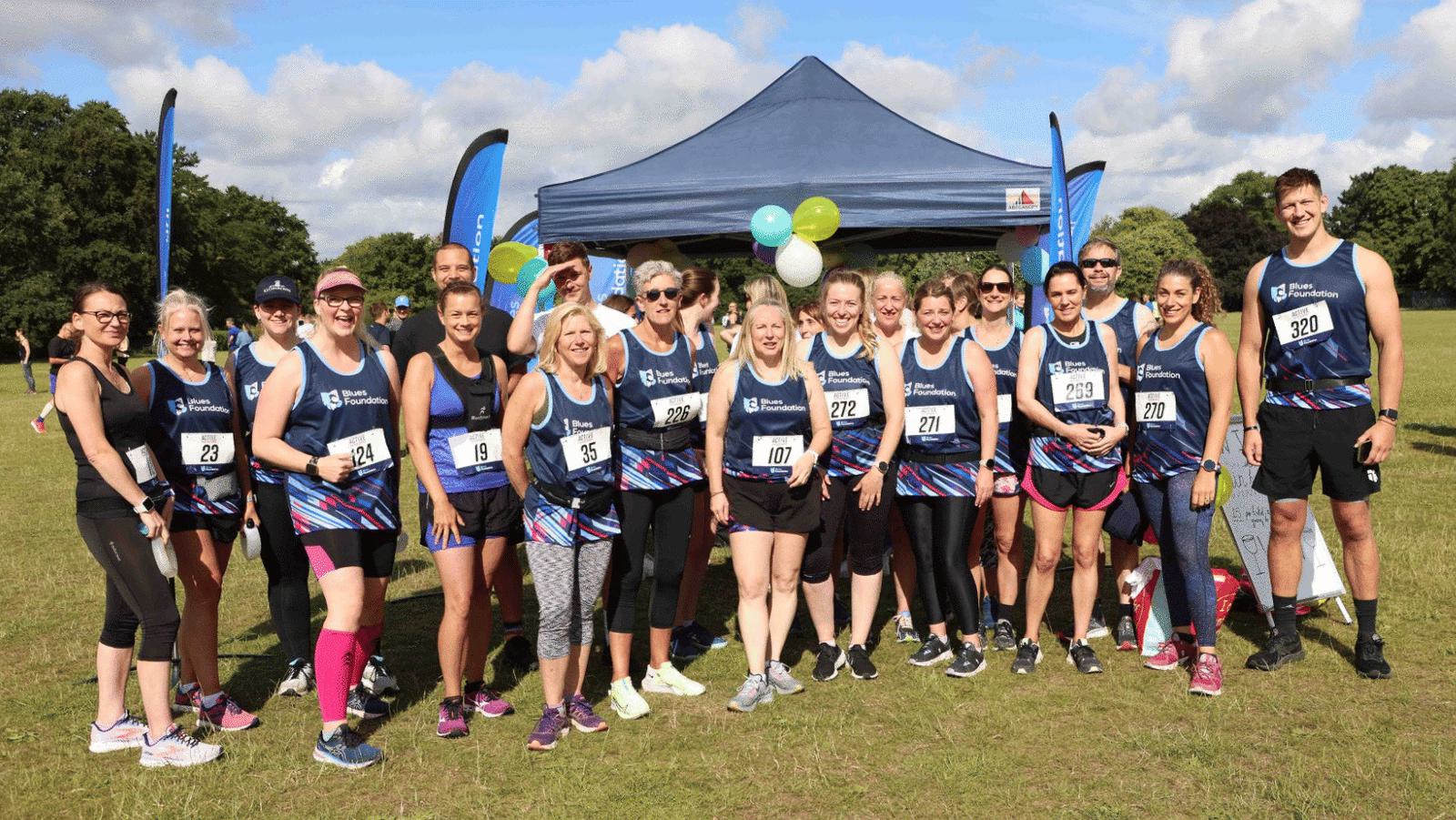 A group of runners in sports attire gather in front of a blue canopy with balloons in a park. They are posing for a photo and wearing race bibs with numbers. Trees can be seen in the background under a partly cloudy sky.