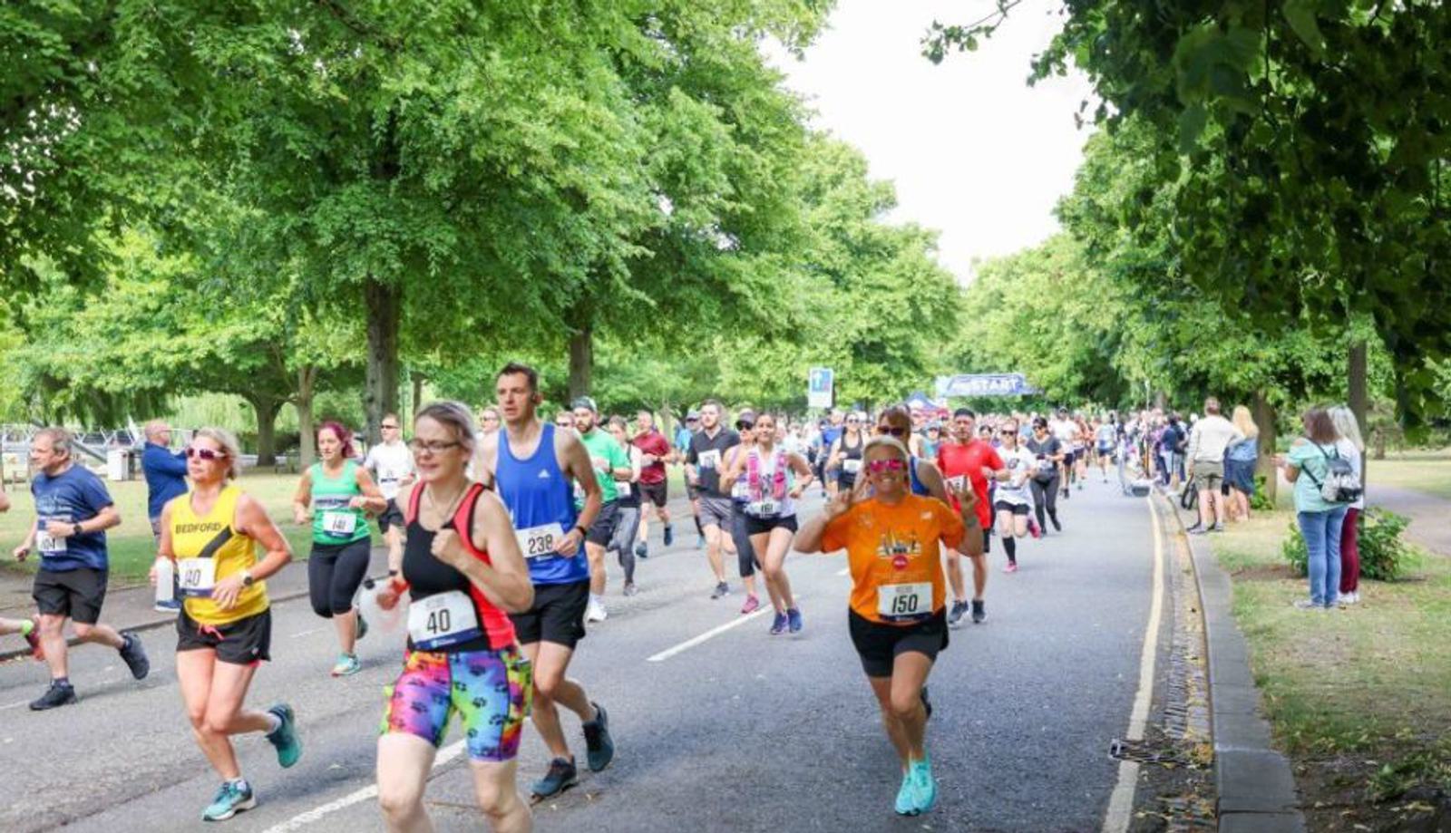 A large group of runners participating in a race on a tree-lined road. Some wear brightly colored outfits and numbered bibs. Spectators can be seen on the sidewalk to the left and right, watching and cheering. The atmosphere appears energetic and lively.