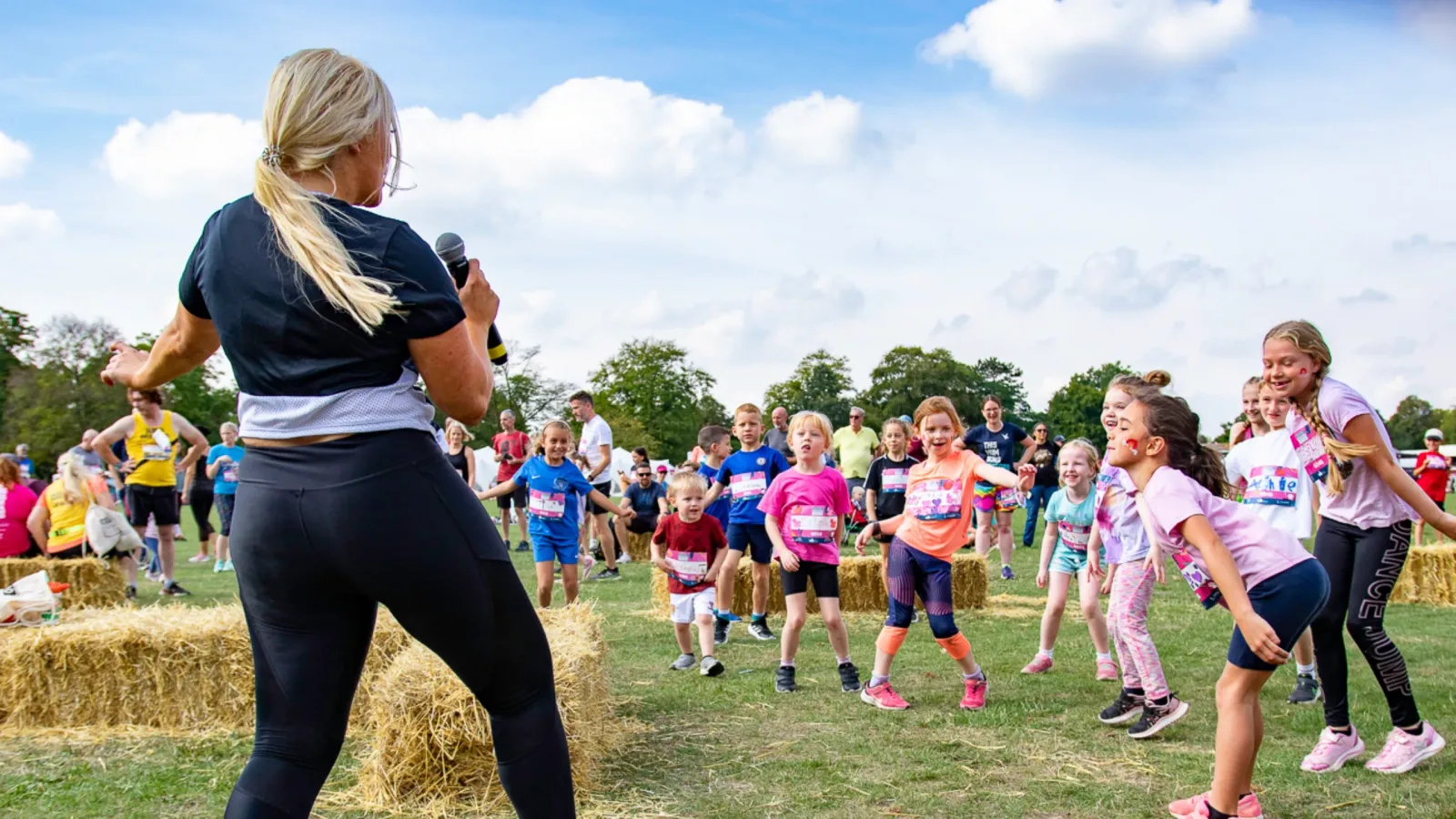A woman, holding a microphone, leads a group of children in an outdoor activity on a sunny day. The kids cheer and participate enthusiastically. The setting includes bales of hay and a crowd of onlookers in a park-like environment with green grass and trees.
