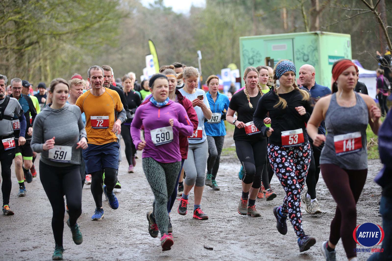 A group of runners participating in an outdoor race on a muddy trail. They are dressed in athletic clothing, with some wearing race bibs and numbers like 591, 72, 590, and 53. Trees and a green portable restroom are visible in the background.