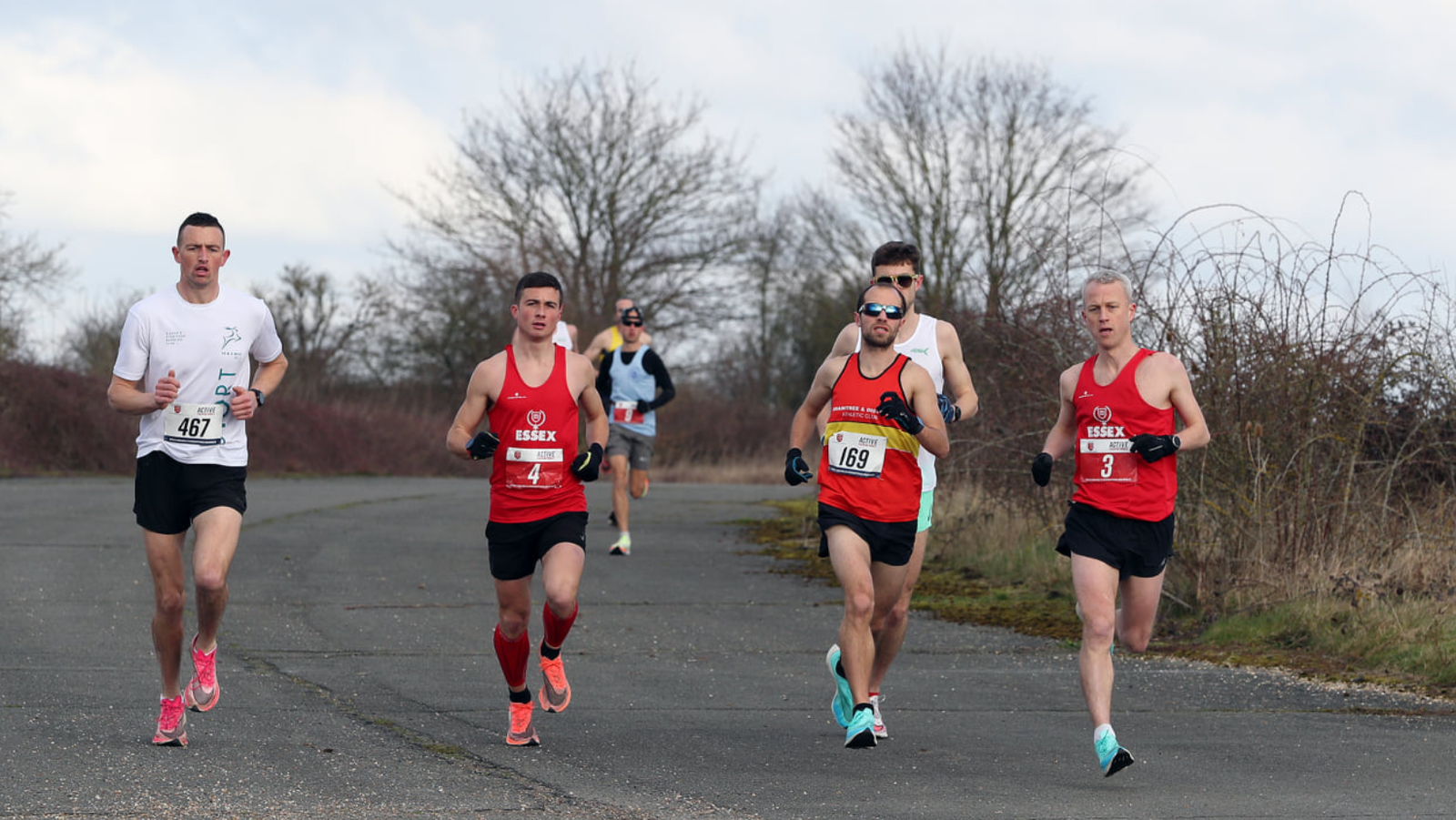 Five runners, three in red tops and two in white tops, race on a paved outdoor path surrounded by leafless trees and bushes. The runners appear focused, with a clear blue sky in the background. One runner wears sunglasses and blue shoes.