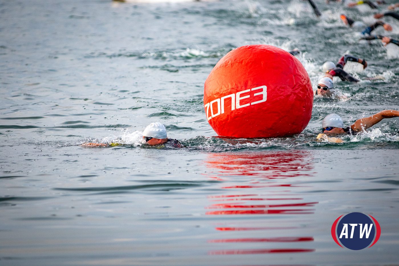 Swimmers in a race are navigating around a large red buoy marked "ZONE3" in the water. The foreground shows two swimmers closest to the buoy, wearing white swim caps and goggles. The background features more racers approaching. The logo "ATW" is visible at the bottom right.