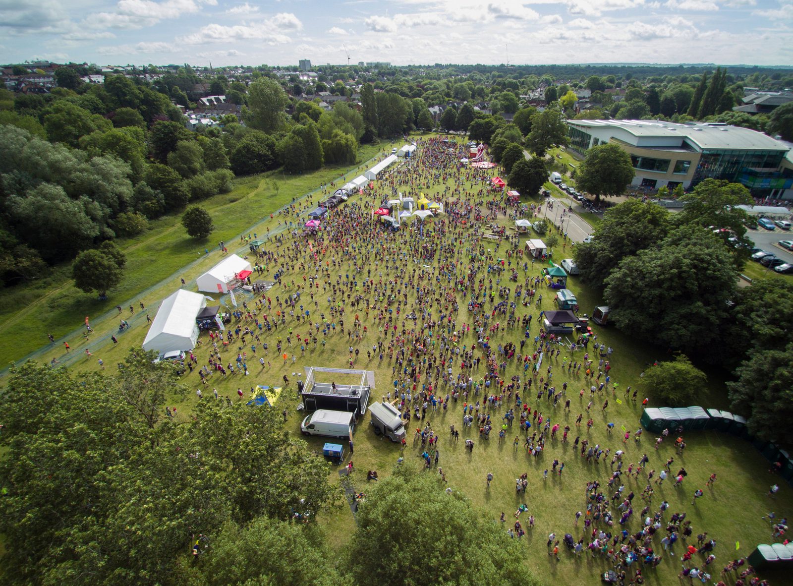 Aerial view of a large outdoor festival in a green park. Crowds of people gather around various tents, booths, and vendor trucks. The park is surrounded by trees and there are several buildings in the background under a partly cloudy sky.