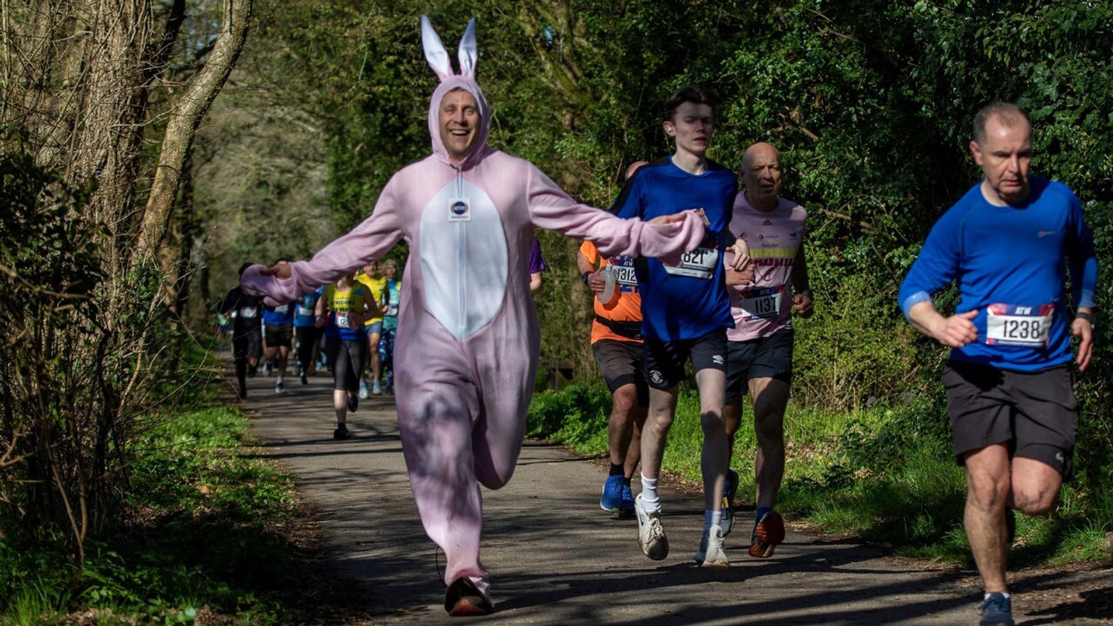 A group of runners participate in a race on a forested path. Among them is a person dressed in a pink bunny costume, smiling with arms outstretched. Other runners are in typical athletic gear, and the path is surrounded by trees and bushes.