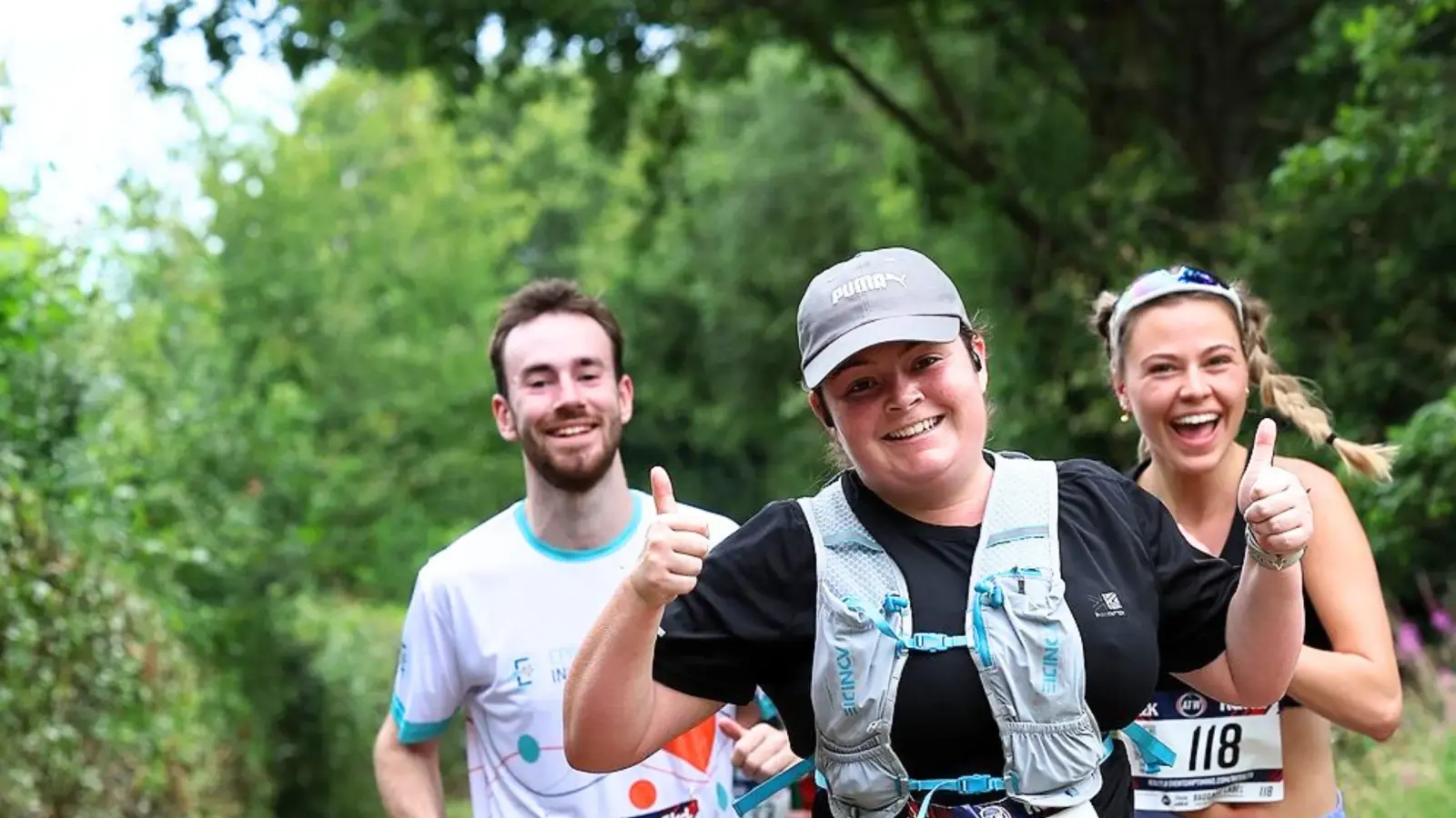 Three runners are smiling and jogging along a tree-lined path. The woman in front is wearing a gray cap and giving thumbs up. Two people behind her, a man in a white shirt and a woman in sunglasses, appear happy and energetic.