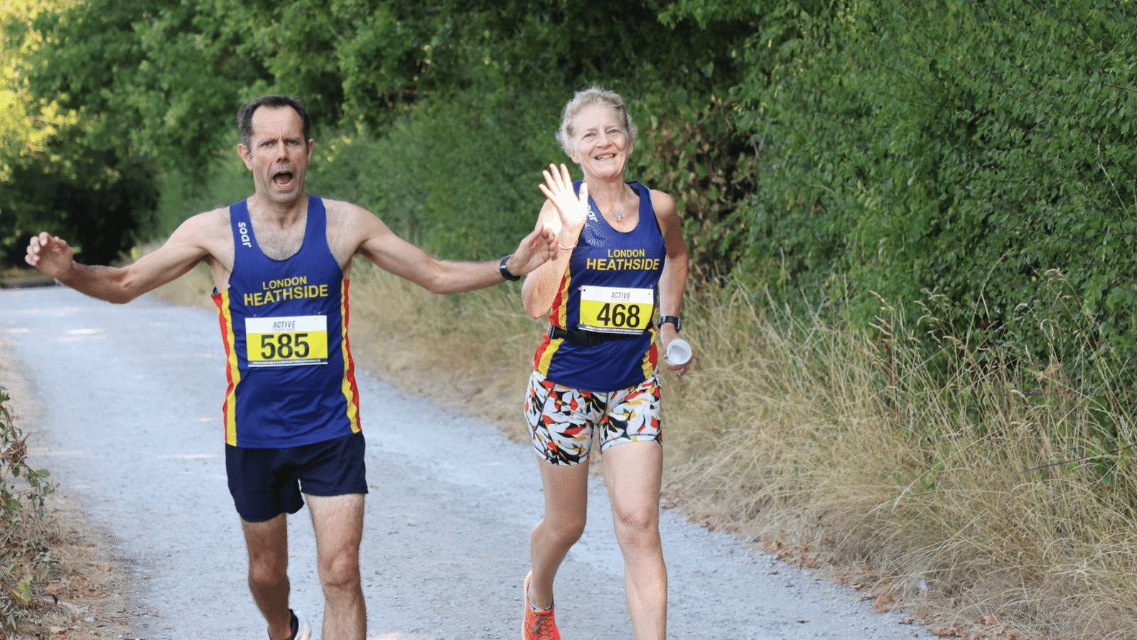 Two runners in "LONDON HEATHSIDE" jerseys are jogging on a gravel path surrounded by greenery. The man (585) looks focused, and the woman (468) is smiling and waving at the camera. Both are wearing race bibs and running gear, enjoying the outdoor activity.