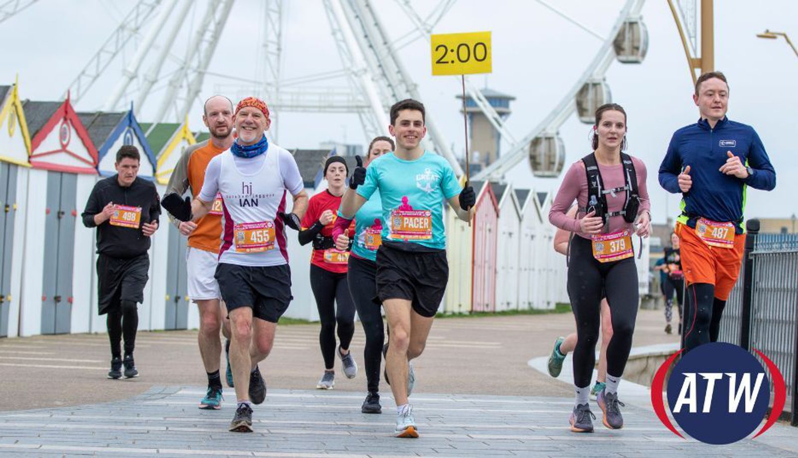 A diverse group of runners participating in a race near a ferris wheel. Some runners wear bibs with numbers, and the time 2:00 is displayed on a sign above. The sky is overcast, and colorful beach huts line the path. The atmosphere is energetic and focused.