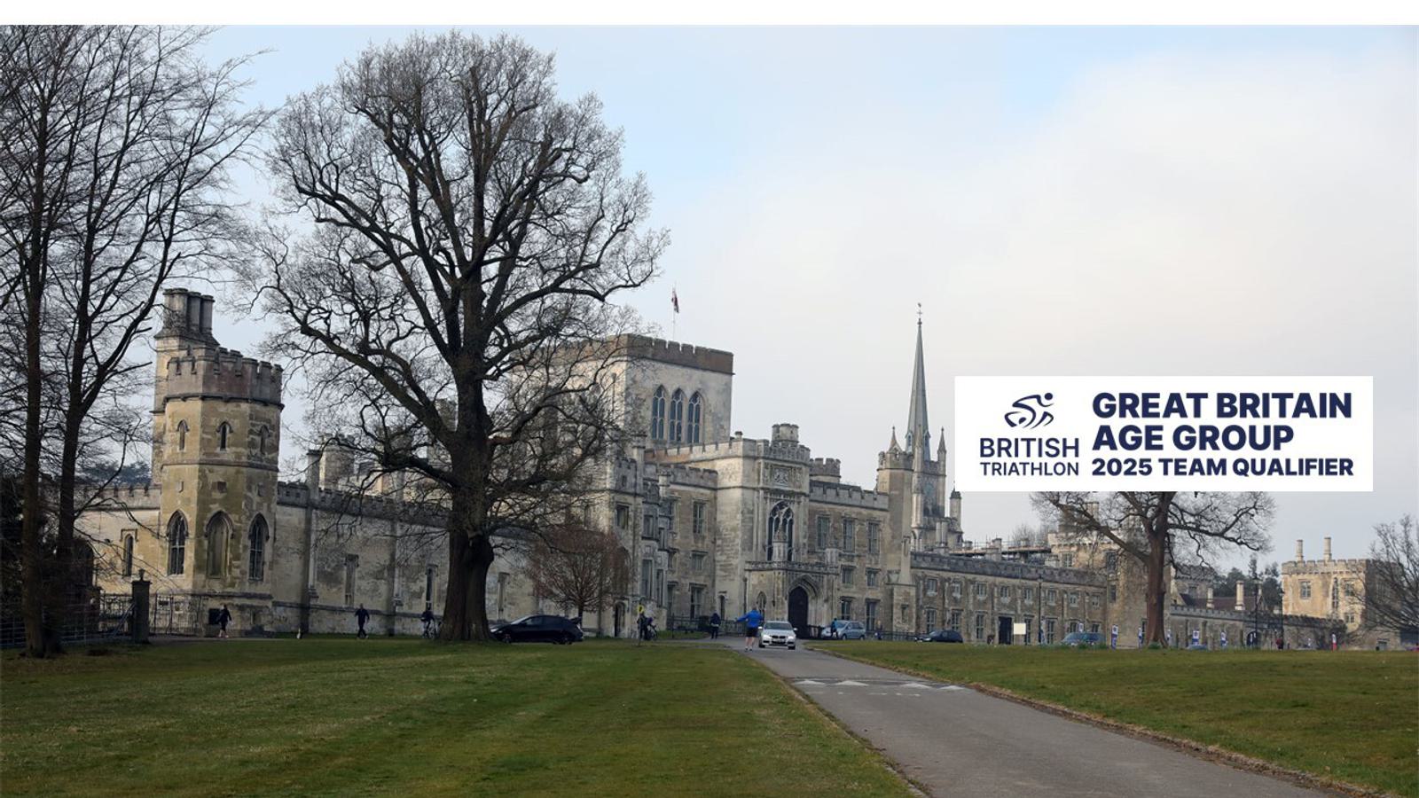 An expansive field leads to a historic stone building complex under a partly cloudy sky. A sign reads "British Triathlon, Great Britain Age Group, 2025 Team Qualifier." Tall trees without leaves stand in the foreground.