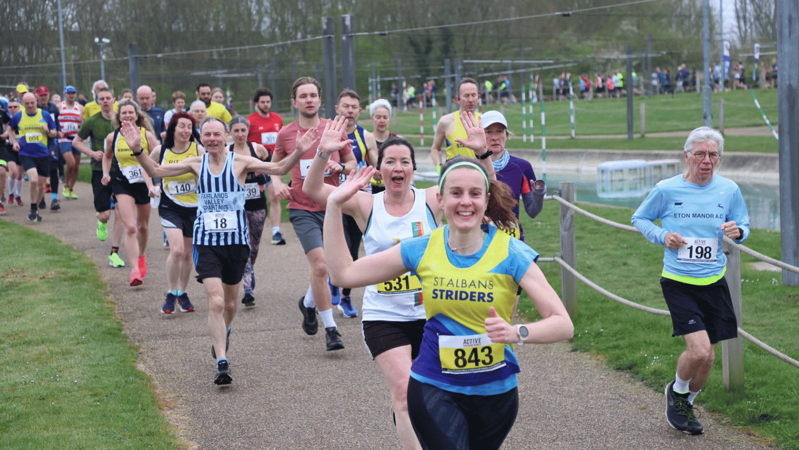 A group of runners participating in a race on a paved path. The runners are wearing various athletic outfits with numbers on their chests. A woman in a yellow "St Albans Striders" shirt with the number 843 is at the front, smiling and waving at the camera.