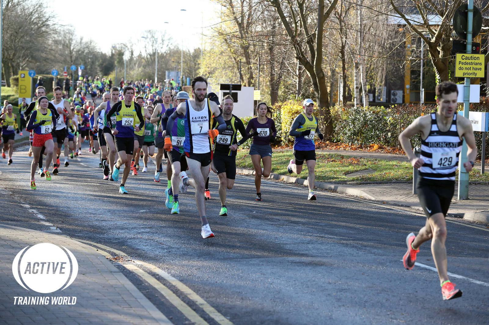 A large group of runners participating in a race on a sunny day. The runners are on a road lined with trees and colorful bushes. One runner is leading the pack, and several runners are wearing brightly colored attire. A sign in the background reads "Caution Pedestrians.