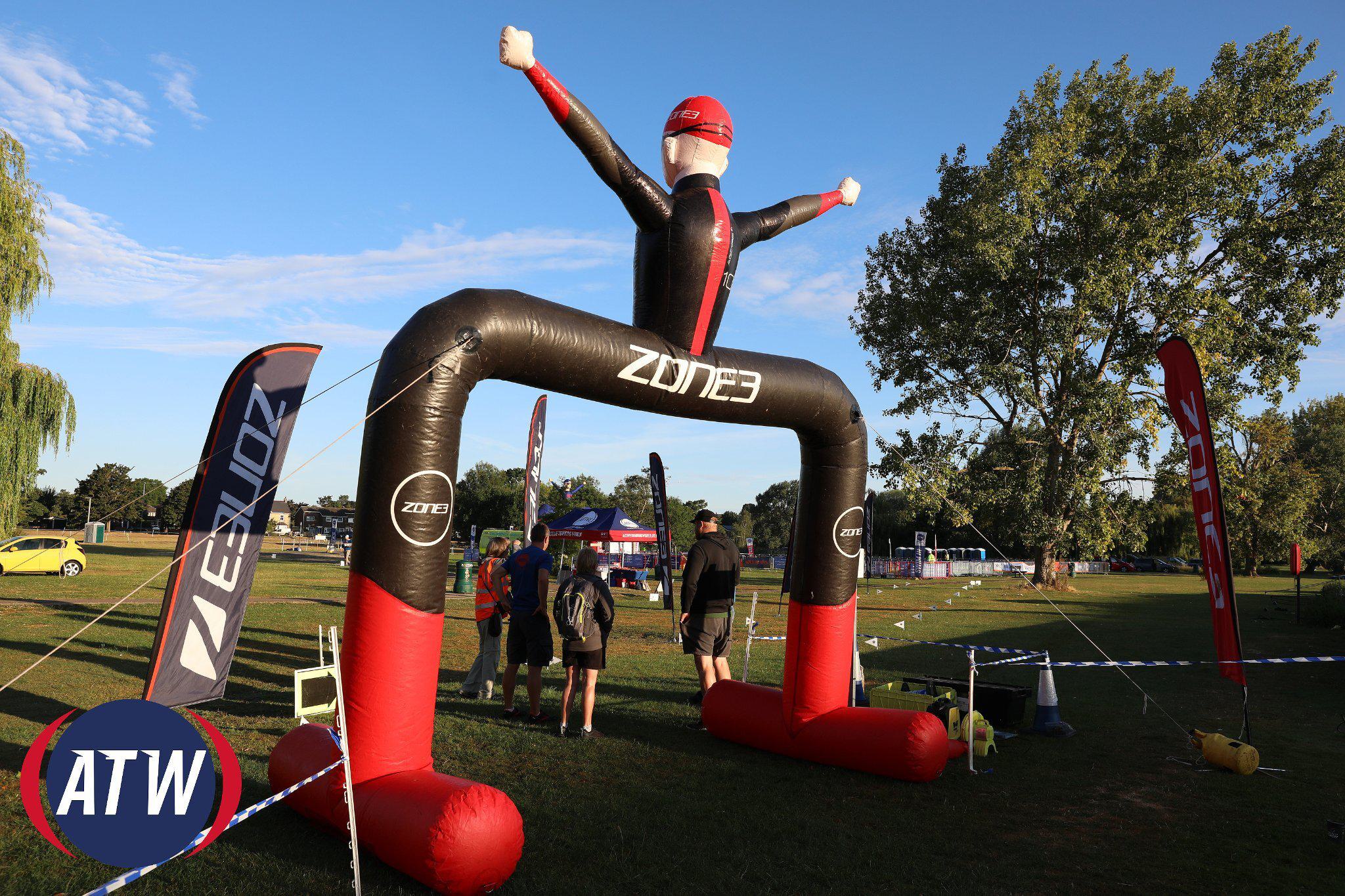 An inflatable arch with a large, inflatable figure stands at the entrance to a competition area. Several people are gathered underneath. The arch is branded with "Zone3." Trees and equipment can be seen in the background. Two flags with logos are on either side.