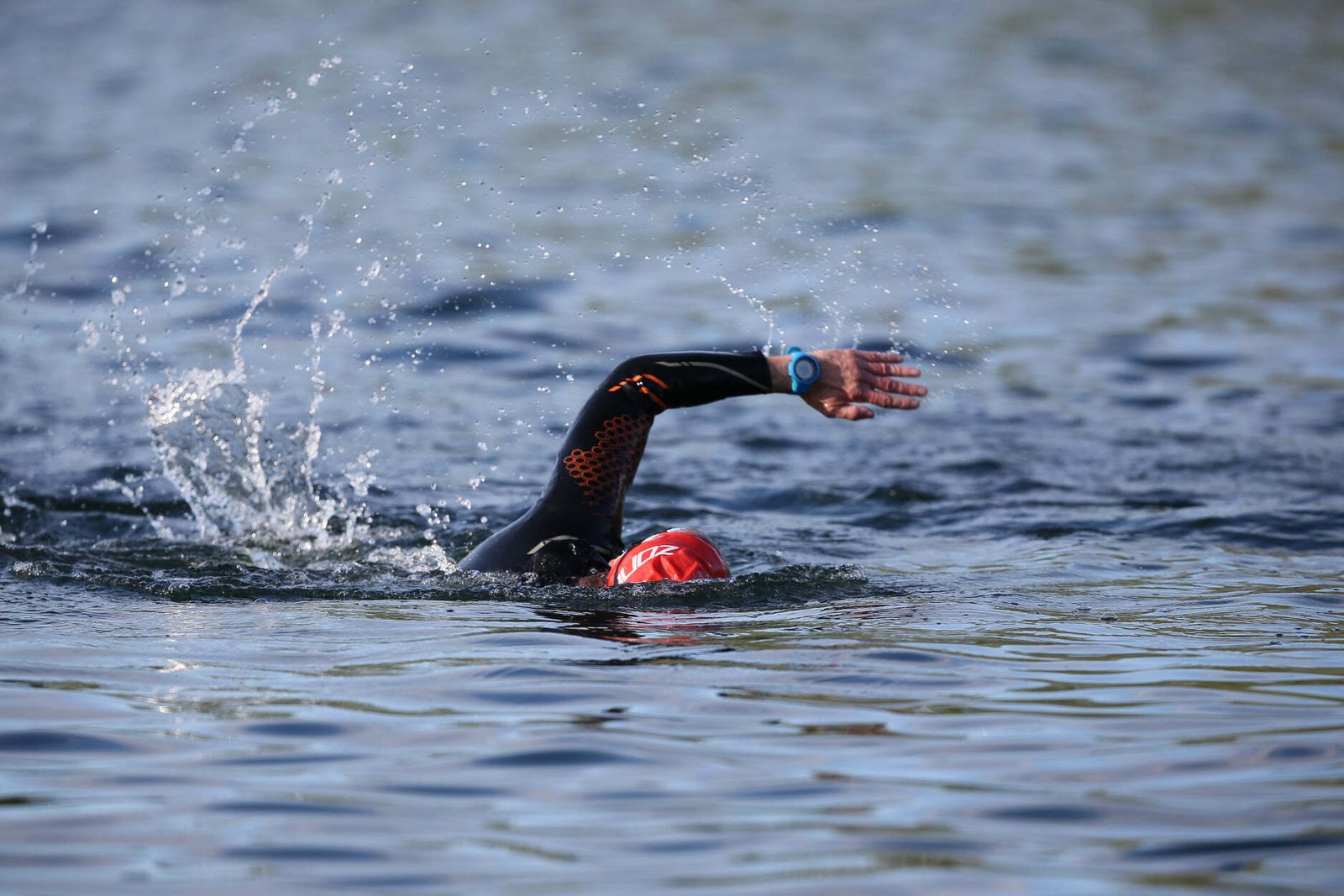 A swimmer wearing a black wetsuit and red swim cap is doing a freestyle stroke in open water. Splashes around the swimmer indicate movement. A light blue wristband is visible on the left wrist. The water surface shows gentle ripples.
