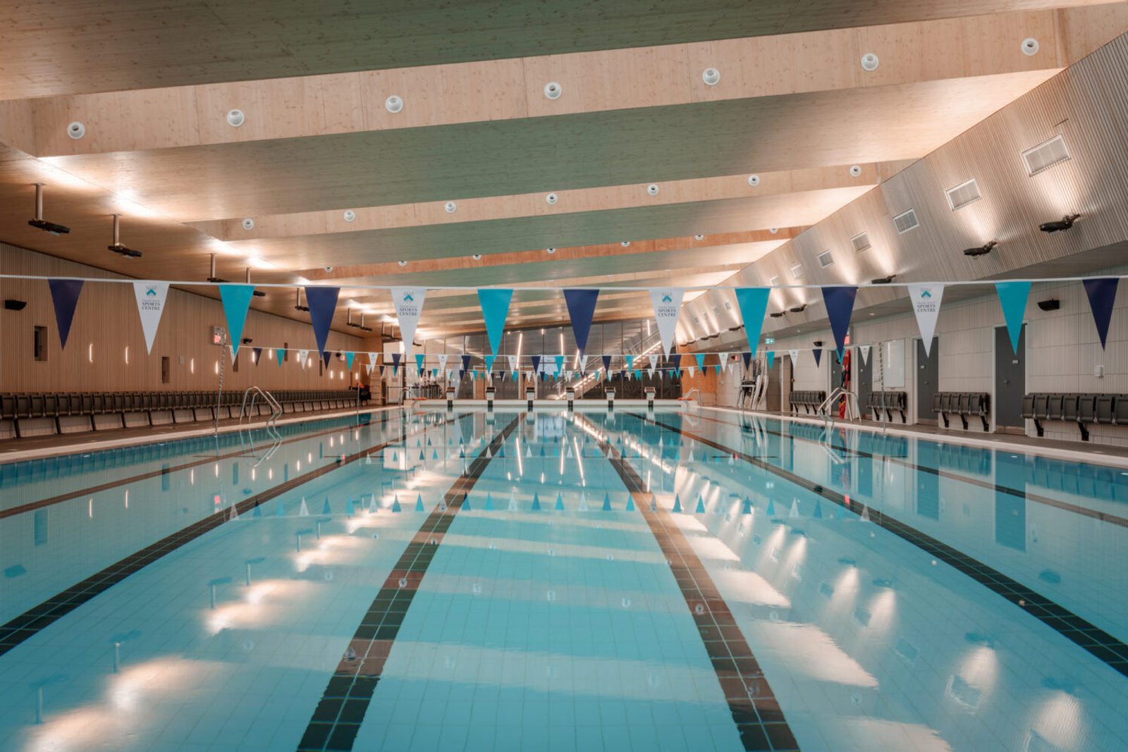 The image shows an empty indoor swimming pool with multiple lanes, decorated with blue and white triangular banners hanging above. The ceiling features wooden panels with overhead lighting. There are seats along the sides of the pool for spectators.