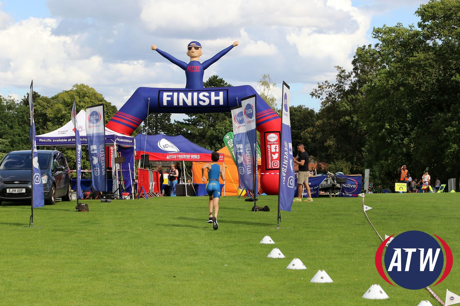 A runner in blue shorts and a blue tank top approaches a blue inflatable finish line arch at an outdoor race event. The arch has "FINISH" written on it and the event includes tents and flags with logos. The grass field is lined with cones, and the sky is partly cloudy.