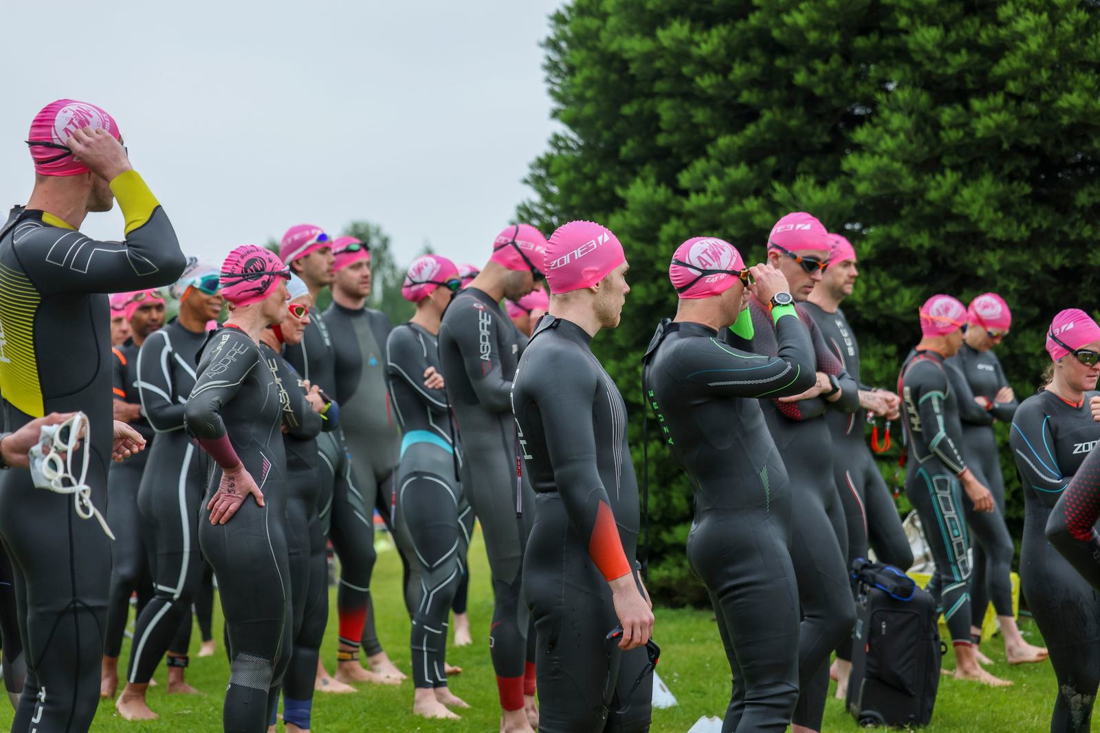 A group of athletes wearing wetsuits and pink swim caps stand on grass, preparing for a swim event. Some adjust goggles and suits. Tall trees form the background under an overcast sky.
