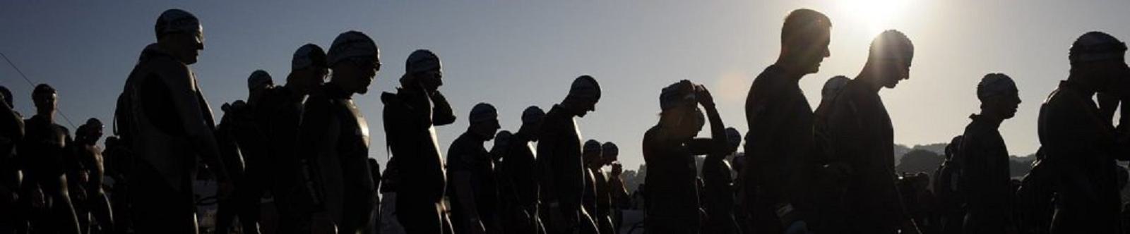 Silhouetted against a sunrise, a group of swimmers wearing swim caps and wetsuits line up in anticipation. The backlighting from the rising sun creates a dramatic effect, casting long shadows and highlighting the anticipation ahead of a swimming event.