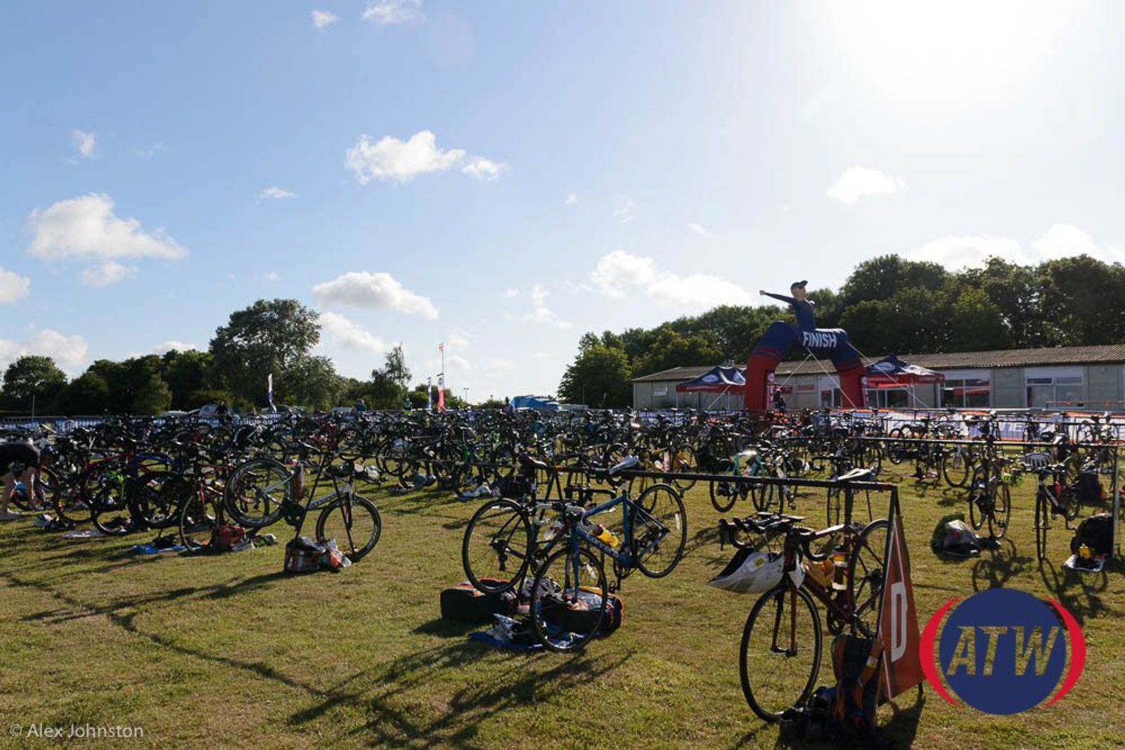 Rows of bicycles are neatly arranged in a grassy triathlon transition area under a clear sky. A colorful inflatable arch marked "FINISH" can be seen in the background, along with a few buildings. The scene is calm, with ample sunlight illuminating the area.