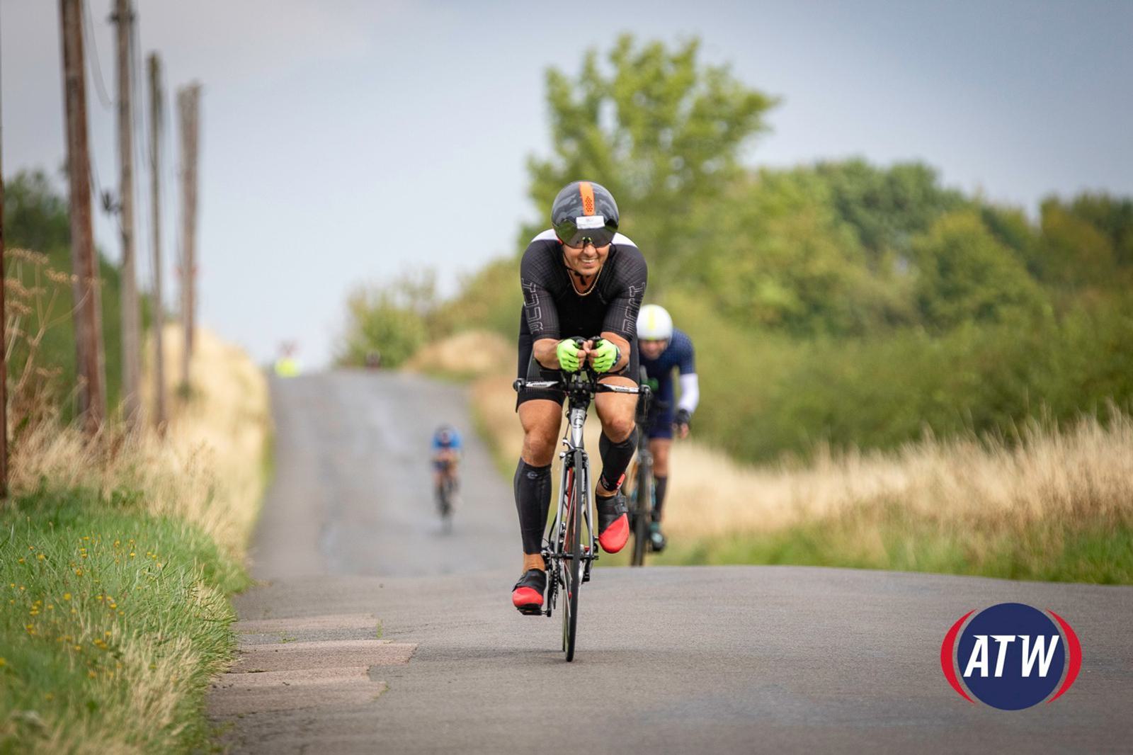 A cyclist wearing a black helmet and sunglasses rides on a country road, closely followed by two other cyclists. They are on a slight incline, with greenery and power poles on either side. The logo "ATW" is visible in the bottom right corner.
