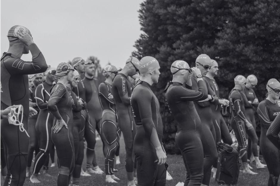 A group of athletes in wetsuits and swim caps stand on grass, preparing to start a triathlon. Some adjust their gear while others look ahead, focused. Large trees are in the background. The image is in black and white.