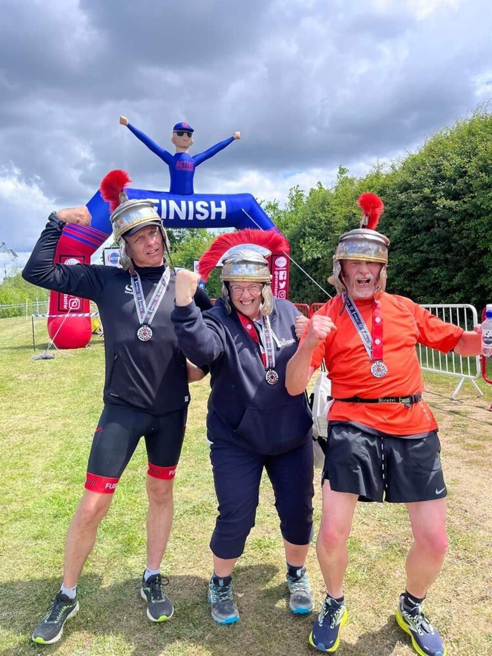Three people in athletic gear and mock Roman helmets celebrate together at a finish line. They are displaying their medals and pointing towards the camera with energetic poses. An inflatable arch marked "Finish" is in the background under a cloudy sky.