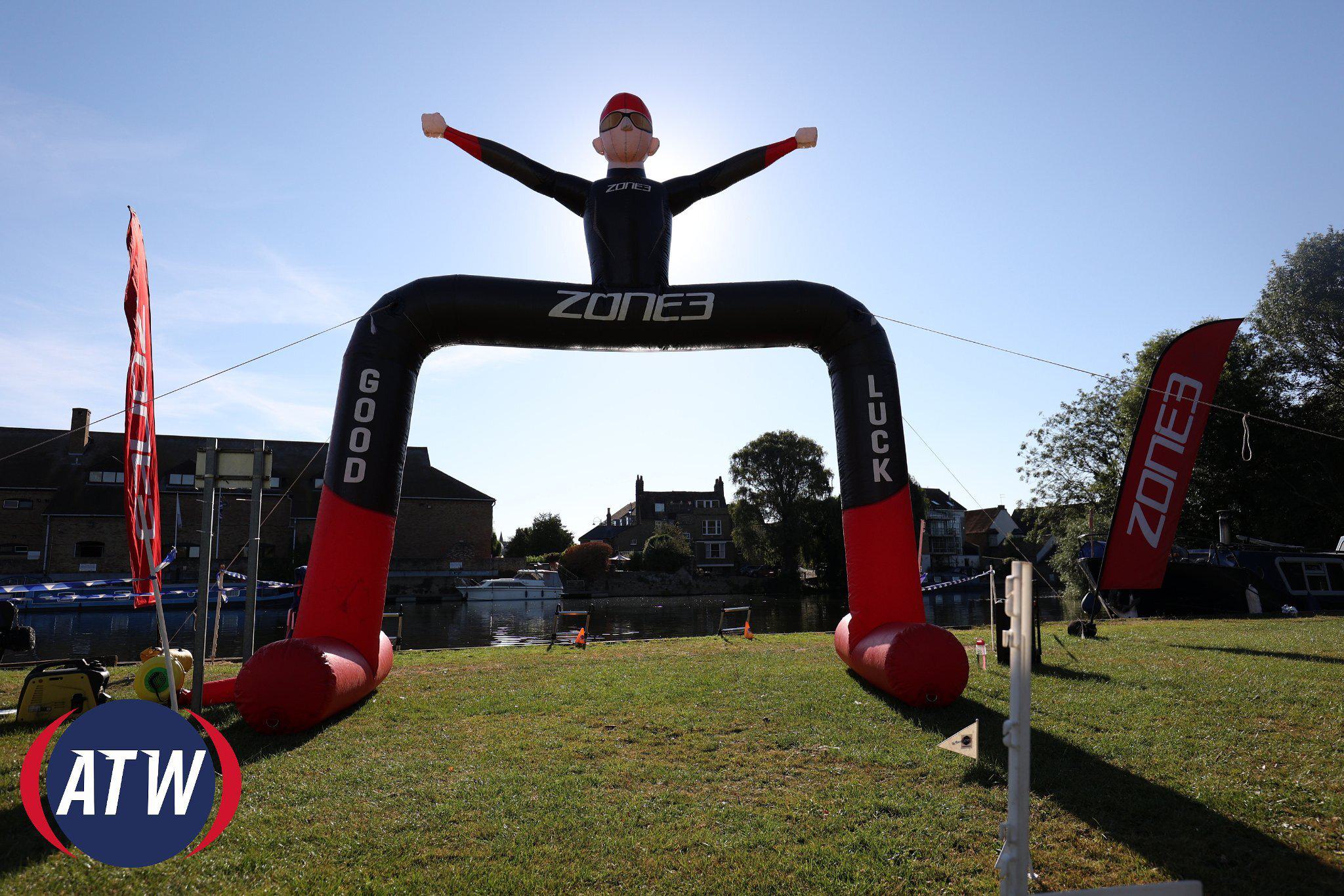 An inflatable figure stands on a grassy area with arms raised, promoting "ZONE3" and displaying "GOOD LUCK" on its legs. There's a body of water and buildings in the background, flags nearby, and the ATW logo visible in the foreground.