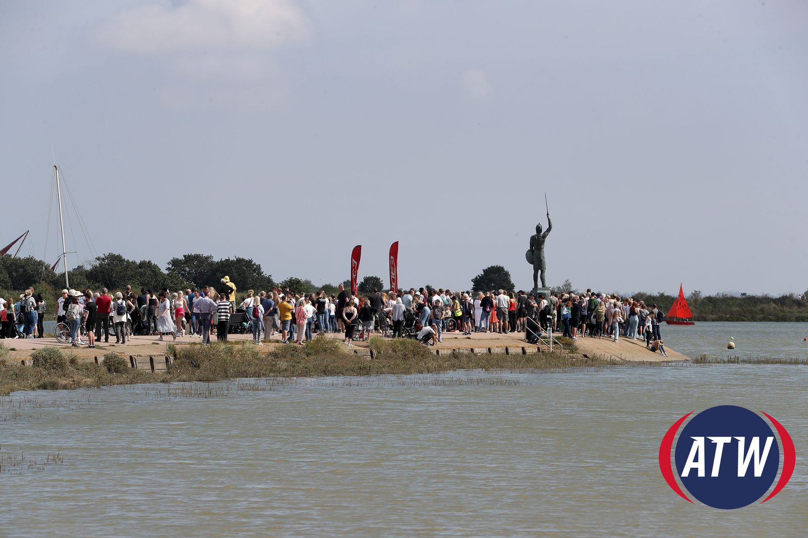 A large crowd gathers on a pier next to the water, with a statue of a person holding a spear in the background. Red flags are placed among the spectators. The sky is partly cloudy, and there is a buoy floating in the water. The ATW logo is in the bottom right corner.