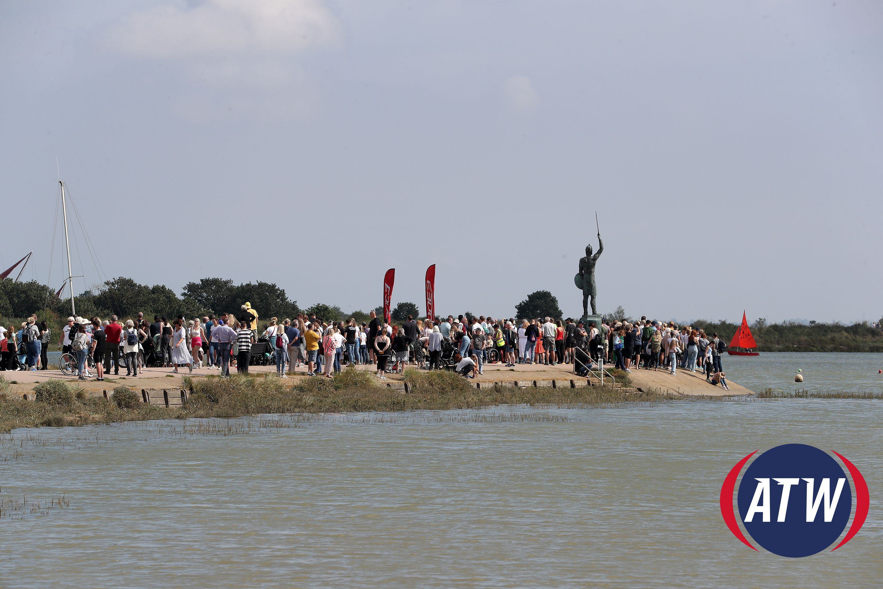 A large crowd gathers on a pier next to the water, with a statue of a person holding a spear in the background. Red flags are placed among the spectators. The sky is partly cloudy, and there is a buoy floating in the water. The ATW logo is in the bottom right corner.