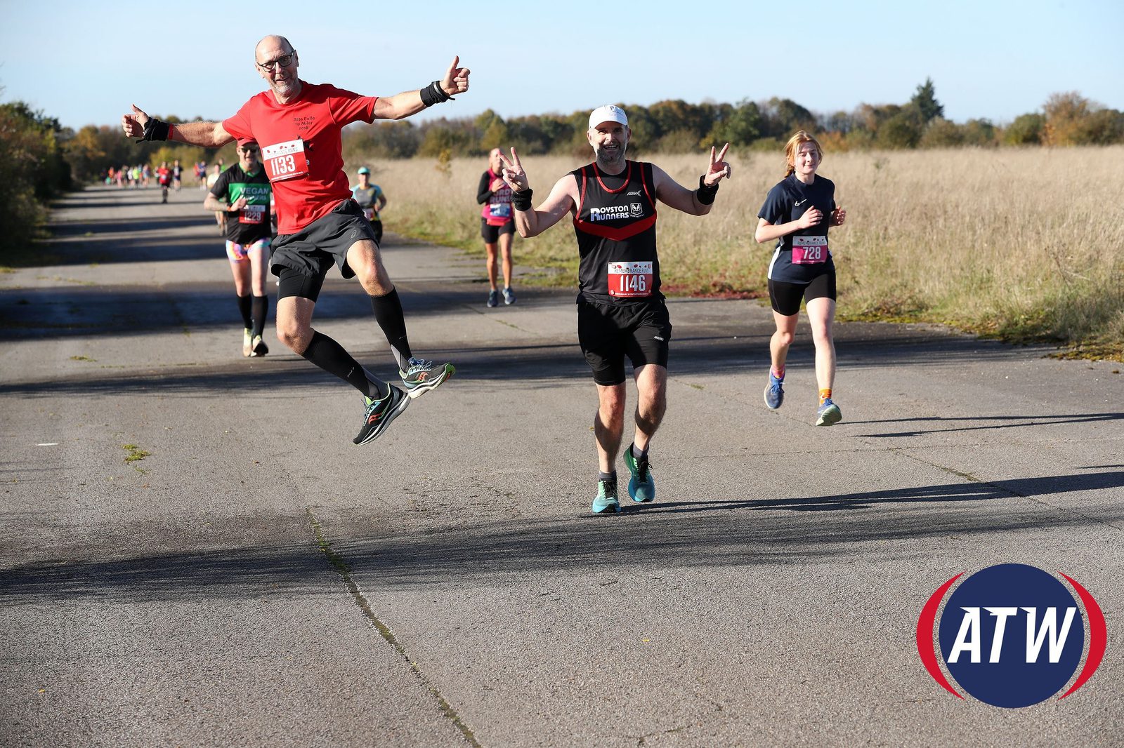 A group of runners participate in a race on a sunny day. In the foreground, a man in a red shirt jumps playfully with a thumbs-up gesture, while others run behind him. The path is flanked by tall grass, and the ATW logo is visible in the lower right corner.