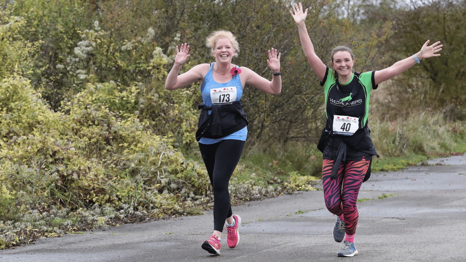 Two female runners, wearing race bibs 173 and 40, smile and raise their arms in celebration while running on a paved path surrounded by greenery. The runner on the left wears a blue tank top, and the one on the right sports a green shirt and zebra-striped leggings.