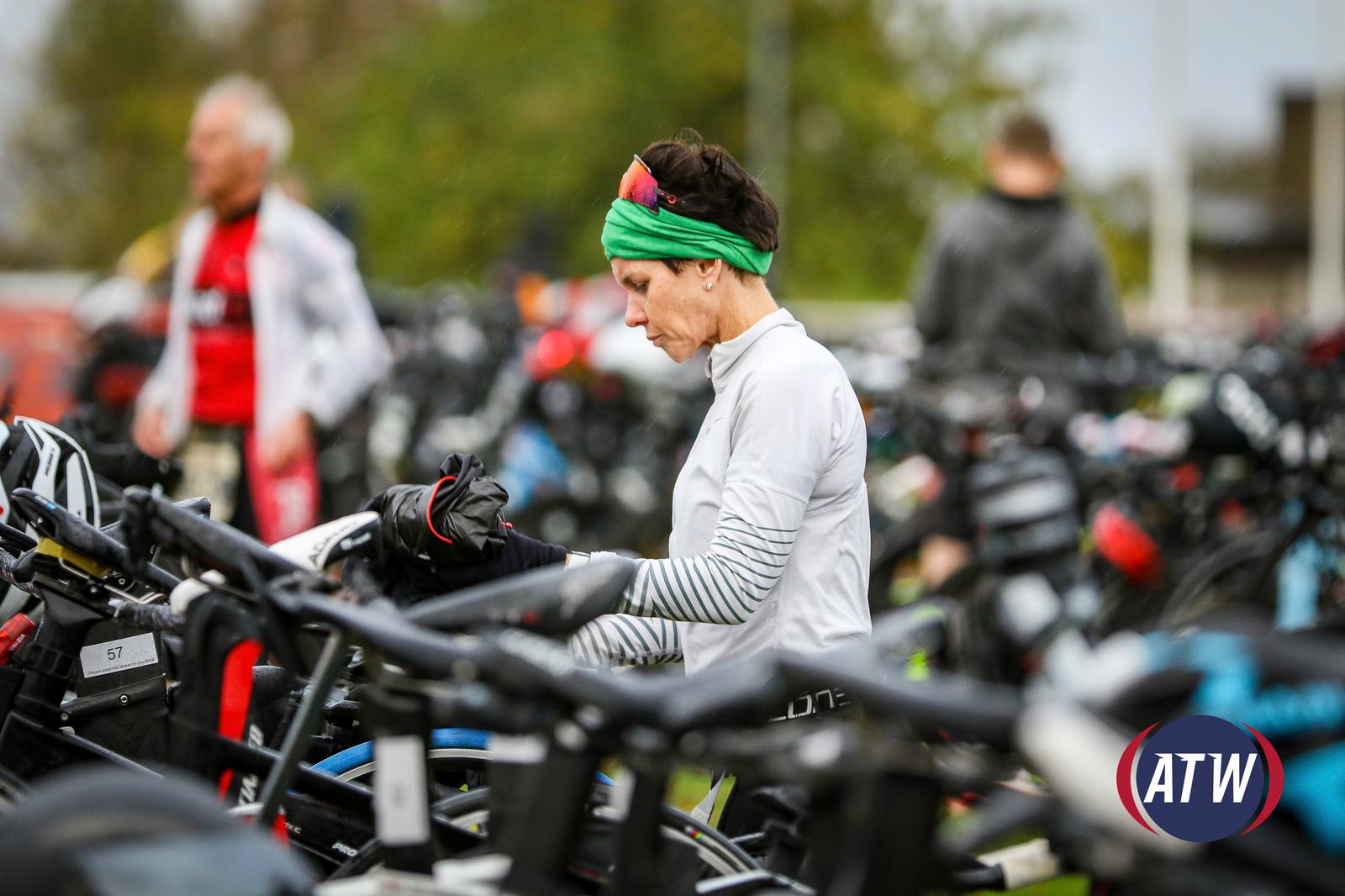 A woman in sports attire adjusts her bike in a crowded transition area filled with bicycles. She wears a green headband and gloves, preparing for a race. Other participants and bikes are in the background, with an ATW logo visible in the corner.