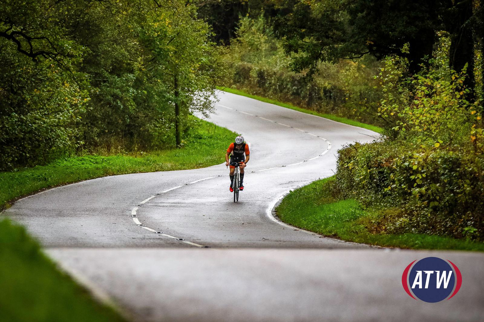 A cyclist wearing a helmet and athletic gear rides along a winding, tree-lined road on a rainy day. The surroundings are lush with greenery, and the wet pavement reflects the overcast sky. A logo with "ATW" is visible in the bottom right corner.
