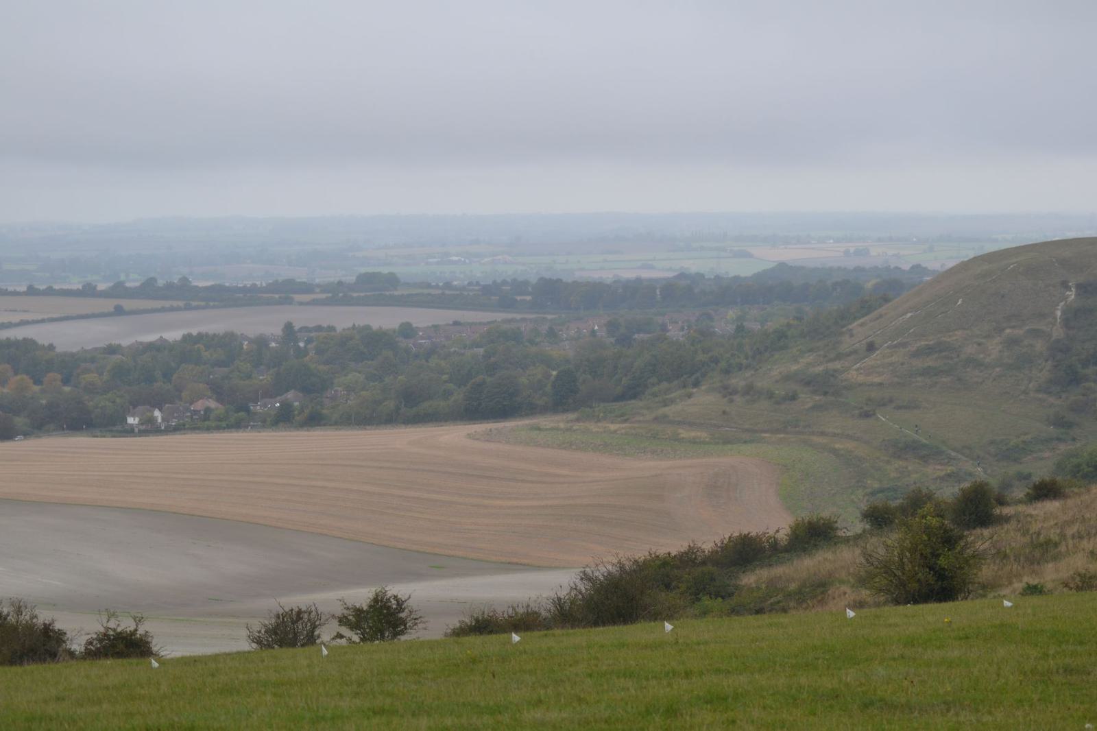 A scenic countryside landscape with rolling hills and farmland. The foreground shows expanses of green grass, while a large, flat, plowed field and patches of trees occupy the middle ground. The background features distant fields and a cloudy sky.