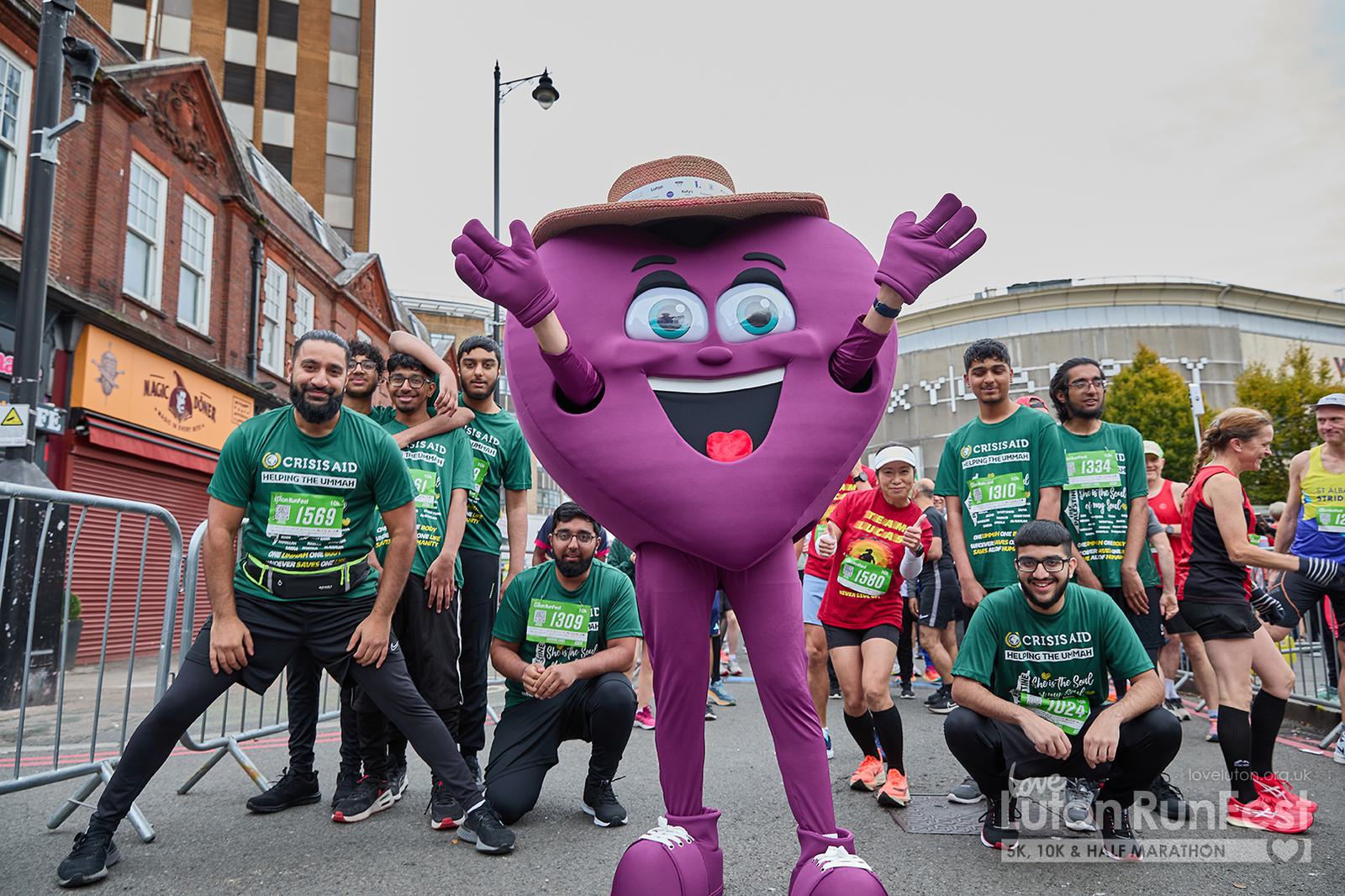 A group of runners in athletic gear pose for a photo with a cheerful purple heart mascot wearing a hat. The setting is a street with colorful buildings and spectators in the background, during a running event.